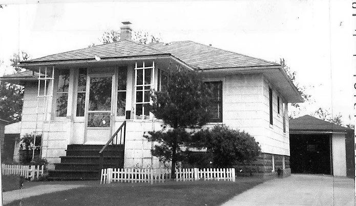 Black-and-white photo of a single-story bungalow with front steps, small picket fence, tree, and driveway leading to a detached garage.