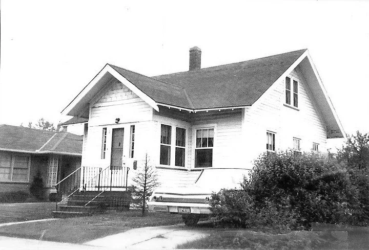Black-and-white photo of a one-and-a-half-story bungalow with front steps and porch, a chimney, shrubs at the right, and a partially visible parked car.