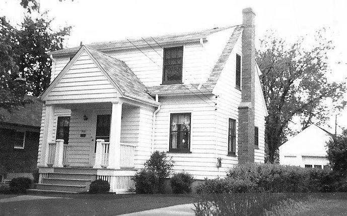 Black-and-white photo of a two-story white clapboard house with a small covered front porch, chimney on the side, driveway, and detached garage in the background.