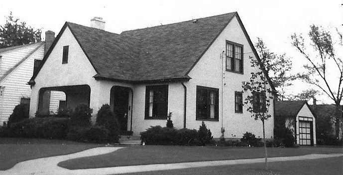Black-and-white photo of a small stucco Tudor-style house with a steep gabled roof, front porch, shrubs along the foundation, and a detached garage to the right.