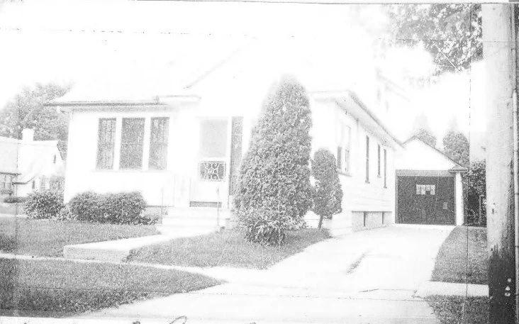 Black-and-white photo of a single-story suburban house with a front lawn, walkway, tall shrub by the entrance, and a driveway leading to a detached garage.