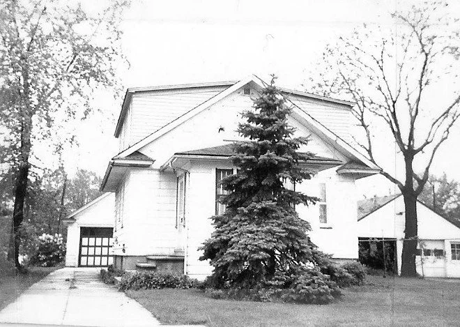 Black-and-white photo of a two-story house with a large evergreen in the front yard, a driveway leading to a detached garage, and trees at the property edges.