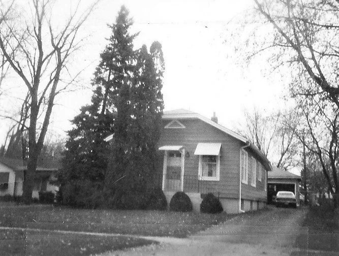 Black-and-white photo of a single-story bungalow with tall evergreen trees in front, a driveway to a detached garage with a car parked inside.