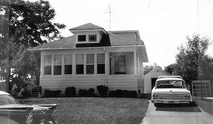 Black-and-white photo of a single-story bungalow with an enclosed front porch and a vintage sedan parked in the driveway.