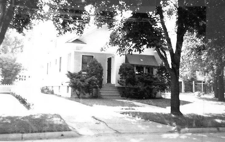 Black-and-white photo of a small house with front steps and shrubs, shaded by large trees along a sidewalk.