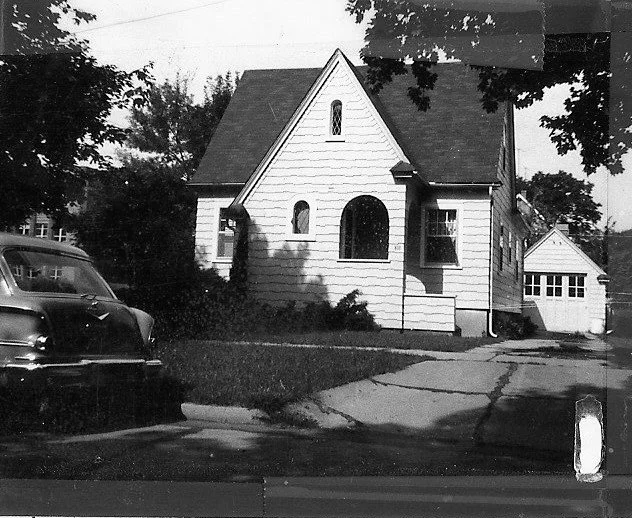 Black-and-white photo of a small gable-front house with an arched entry, driveway leading to a detached garage, and a vintage car parked at the curb.