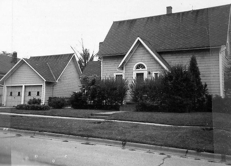 Black-and-white photo of a residential house with a steep gabled roof, arched front window, dense shrubs across the front lawn, and a detached two-car garage beside a sidewalk.