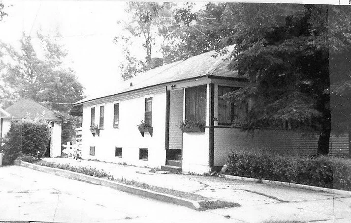 Black-and-white photo of a single-story bungalow with a small screened porch, window flower boxes, driveway curb in front, and trees along the side.