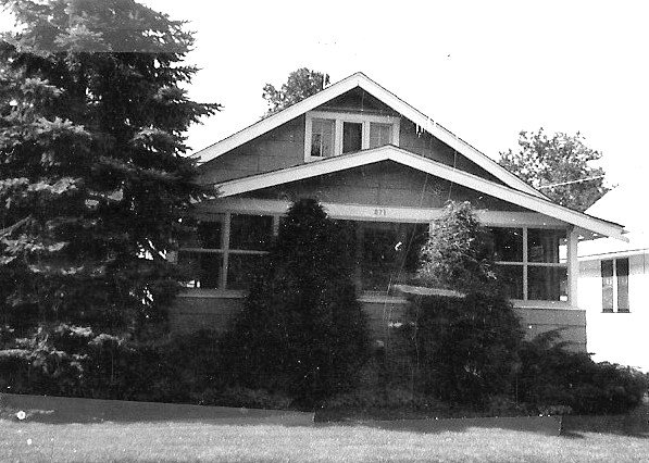 Black-and-white photo of a bungalow-style house with a covered front porch partly obscured by tall evergreens and shrubs.