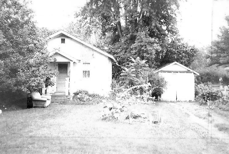 Black-and-white photo of a small wood-frame house with a front porch, lawn and garden bed, and a detached garage to the right, surrounded by trees.