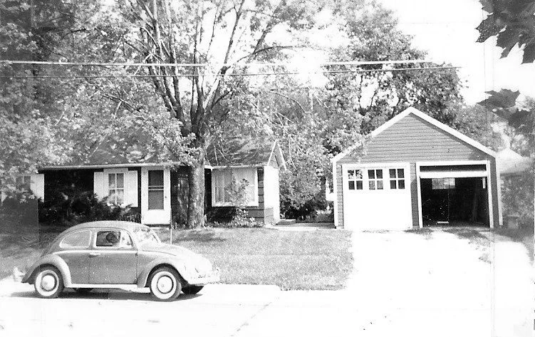 Black-and-white photo of a single-story midcentury house with a detached garage and a Volkswagen Beetle parked on the street in front.