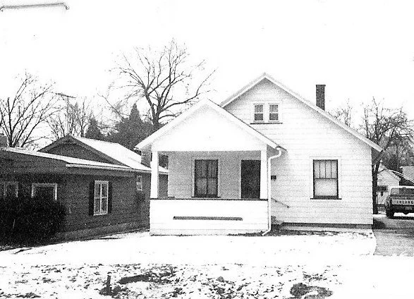 Small white bungalow with a covered front porch and light snow on the yard, neighboring house to the left.