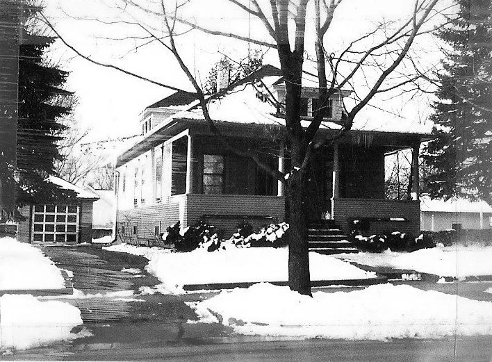 Black-and-white photo of a bungalow-style house with a covered front porch, a large bare tree in the yard, snow-covered lawn and driveway leading to a detached garage.