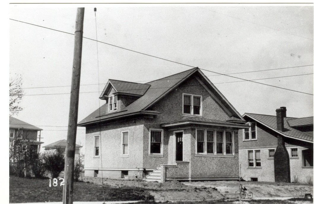 Black-and-white photo of a two-story stucco house with a small front porch, utility pole and overhead wires in the foreground, and neighboring houses visible.
