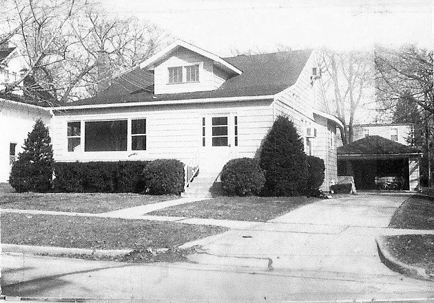 Black-and-white photo of a single-story bungalow with a front porch, trimmed shrubs, driveway, and detached carport.