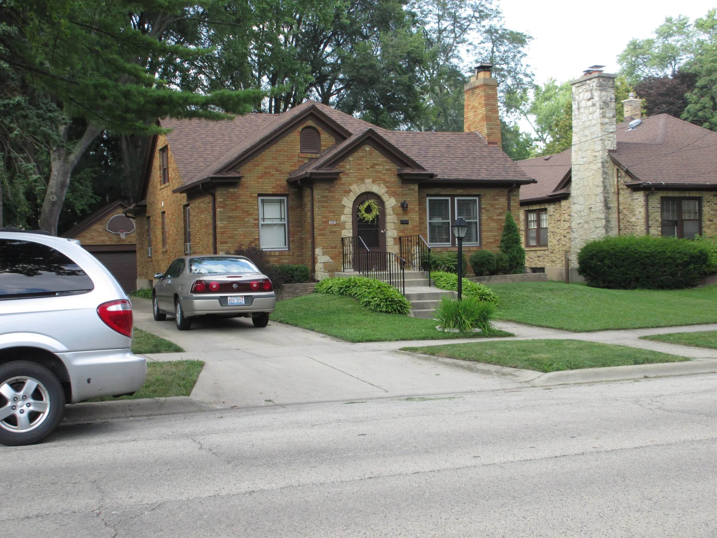 Brick bungalow with an arched front door, front steps and small lawn, two cars parked in the driveway and trees behind the house.