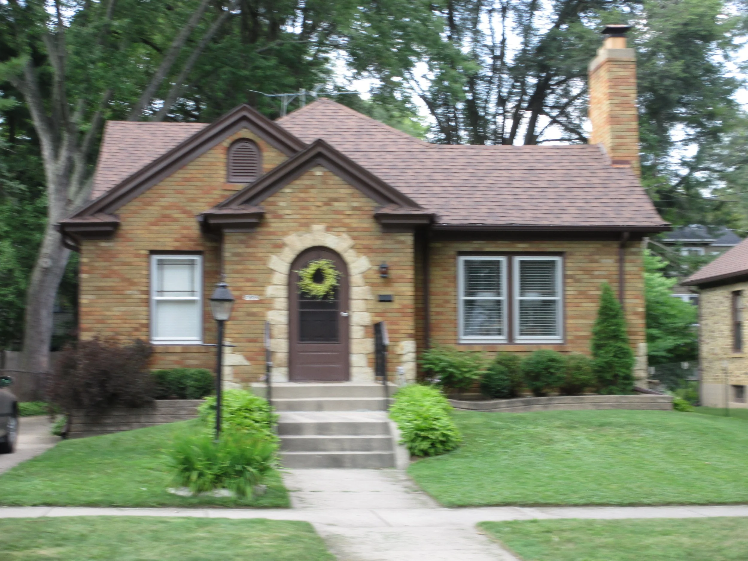 Small brick cottage front with peaked roof, central arched door with a wreath, two windows, front steps, and a chimney surrounded by trees and lawn.