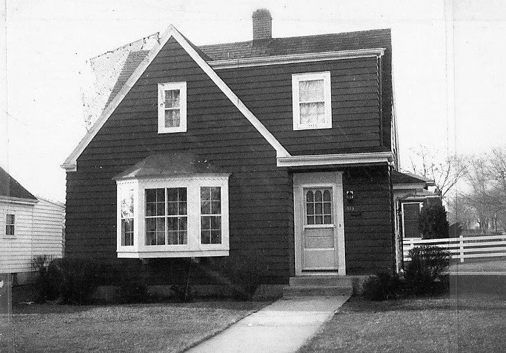Black-and-white photo of a two-story gable-front house with a bay window, front door, and a sidewalk leading to the entrance.