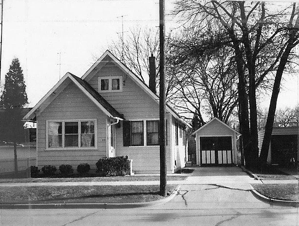 Black-and-white photo of a small bungalow-style house with a front porch, driveway leading to a detached garage, bare trees and a utility pole.