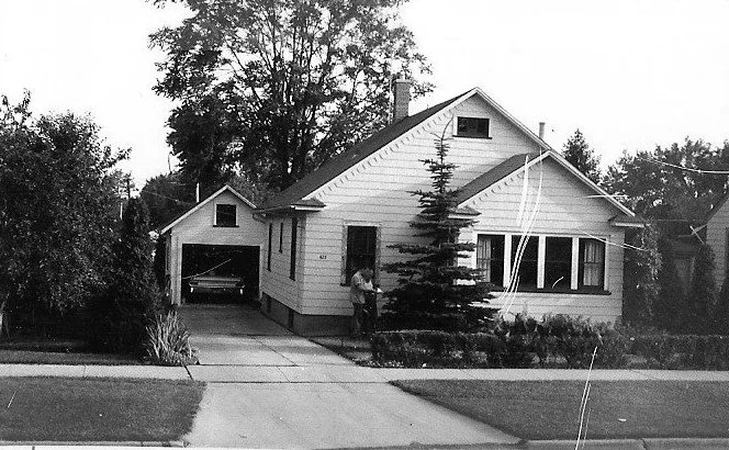 Black-and-white photo of a one-story house with front lawn, driveway to a detached garage, a person at the front door, and a car visible inside the garage.
