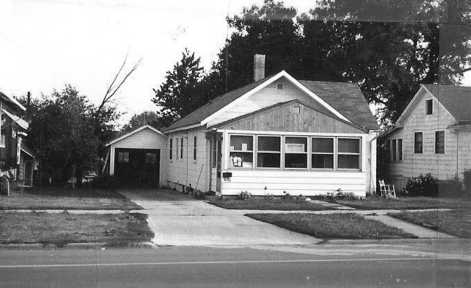 Black-and-white photo of a single-story bungalow with an enclosed front porch, concrete driveway leading to a detached garage, and neighboring houses.
