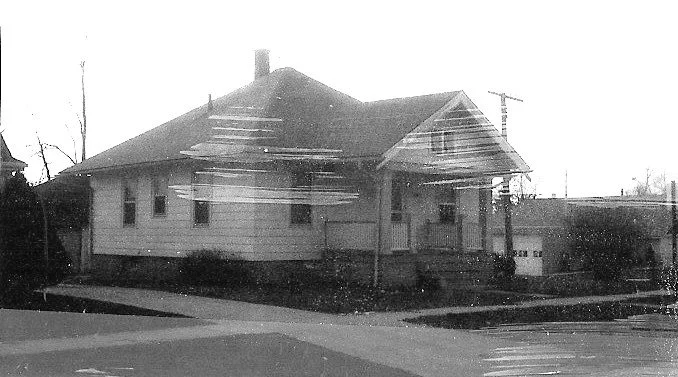 Black-and-white photo of a single-story house with a covered front porch at a street corner, driveway and garage visible to the right.