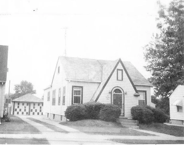 Black-and-white photo of a small single-story house with a steep front gable and arched doorway, shrubs in front, and a driveway leading to a detached garage.
