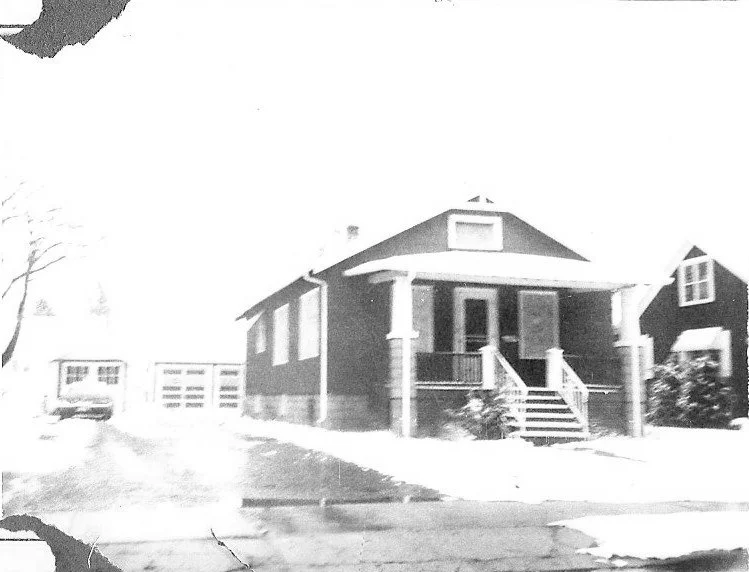 Black-and-white photo of a small single-story house with a front porch and steps, a snowy yard and driveway leading to a garage with a car.