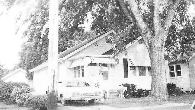 Black-and-white photo of a small bungalow with a large front tree, a car parked in the driveway and a person bending near the shrubs.