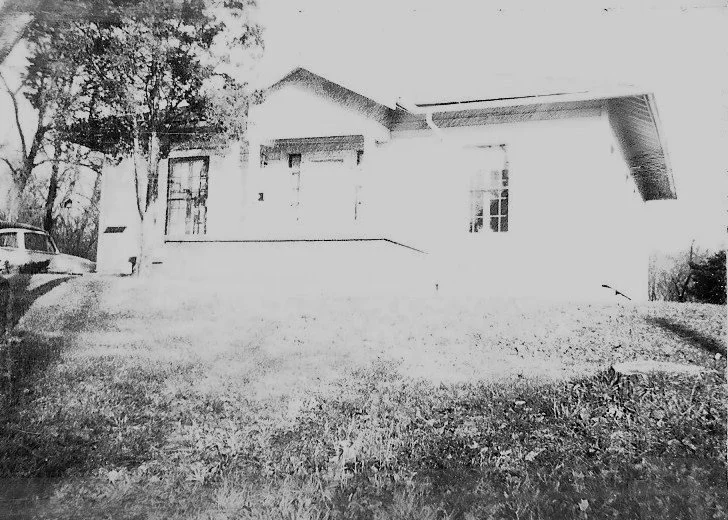 Black-and-white photo of a single-story bungalow with a small front porch, grassy lawn, trees at left and a parked car partially visible.