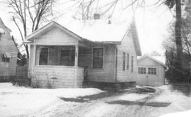 Black-and-white photo of a small single-story house with a covered front porch and a detached garage, snow on the ground and bare trees in the yard.