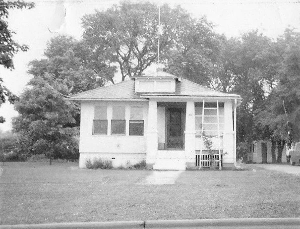 Small one-story bungalow with an enclosed front porch, central entry steps, and large trees in the yard behind.