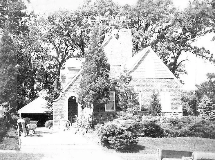 Black-and-white photo of a brick cottage with steep gables and surrounding trees; a person pushing a wheelbarrow on the driveway toward a detached garage.