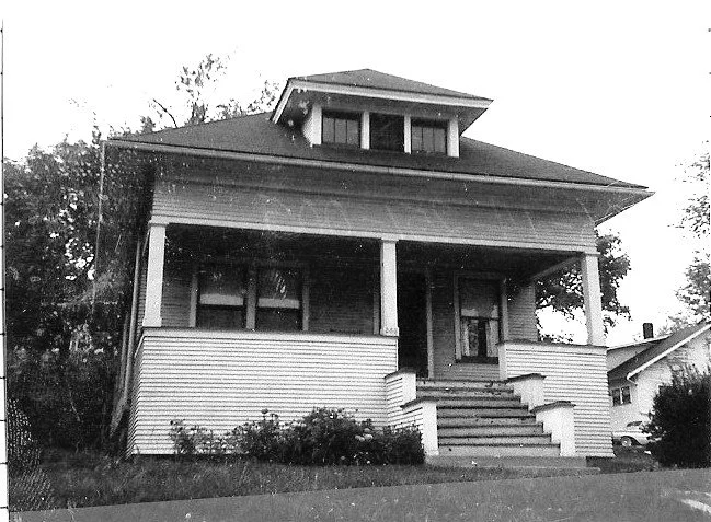 Black-and-white photo of a two-story Craftsman-style bungalow with a wide covered front porch, central steps, and a dormer window.