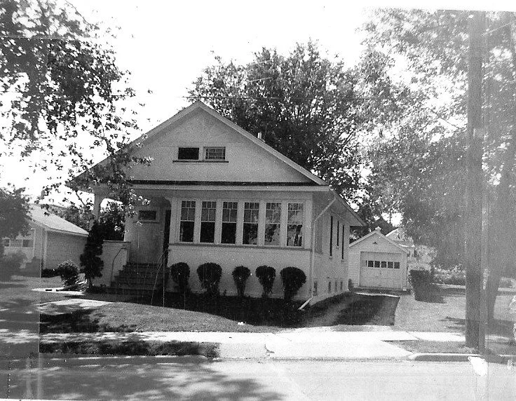Black-and-white photo of a single-story bungalow with an enclosed front porch, row of trimmed shrubs, driveway to a detached garage, and large shade trees.