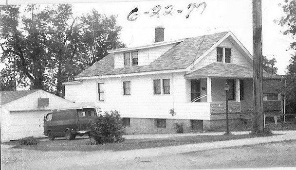 Black-and-white photo of a two-story wood-clad house with a front porch, a van parked beside a detached garage, and "6-22-77" handwritten at the top.