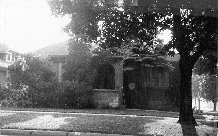 Black-and-white photo of a tree-shaded brick bungalow with an arched front porch, shrubs, and a lawn-lined sidewalk. (Copy)