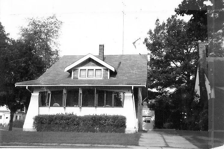 Black-and-white photo of a single-story bungalow with enclosed front porch, low shrubs, driveway to the right, and large trees in the background.