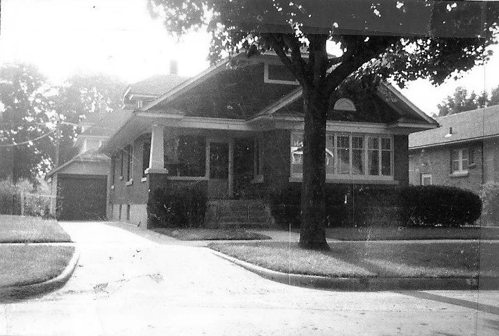 Black-and-white photo of a one-story brick bungalow with a covered front porch, large tree in the front yard, and a driveway leading to a detached garage.