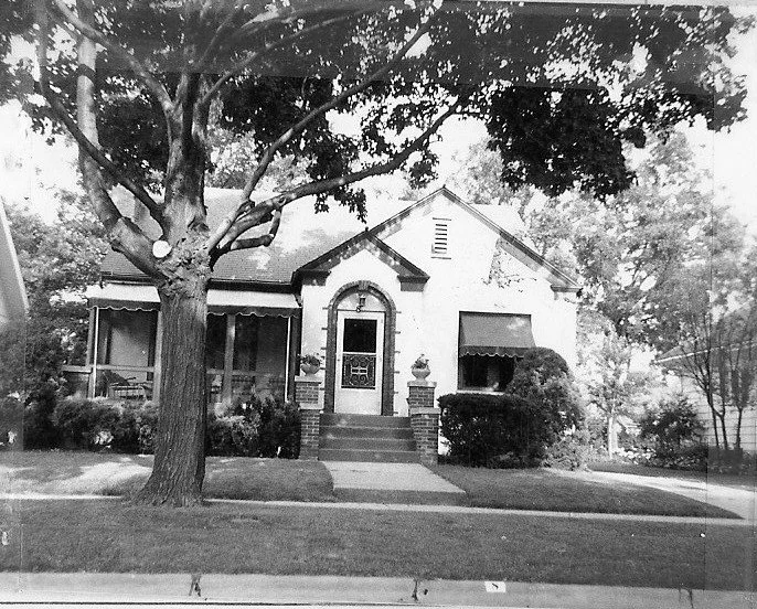 Black-and-white photo of a small bungalow with a front porch, arched doorway, large tree, and manicured lawn.