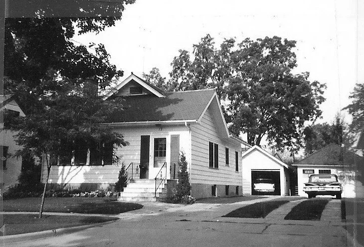 Black-and-white photo of a single-story clapboard house with front steps and a small porch, a detached garage and two cars in the driveway, shaded by large trees.