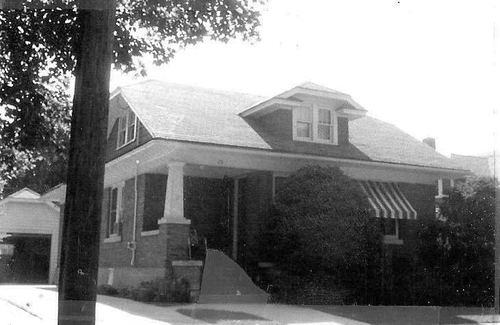 Black-and-white photo of a single-story brick bungalow with a covered front porch, striped window awning, large front shrub, and detached garage.