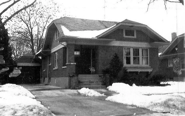 One-story brick bungalow with a snow-covered yard and driveway, bare trees and a detached garage in the background.