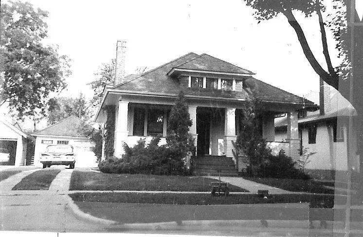 Black-and-white photo of a one-story bungalow with a front porch, shrubs and tall trees, a driveway with a parked car, and a detached garage.