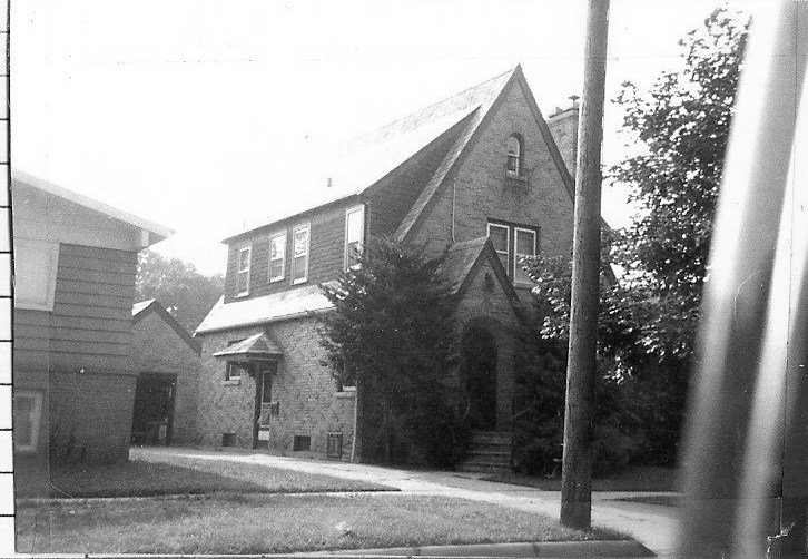 Black-and-white photo of a two-story stone house with a steep gabled roof, arched front entrance and steps, flanked by trees, a driveway and nearby garage, with a telephone pole in the foreground.