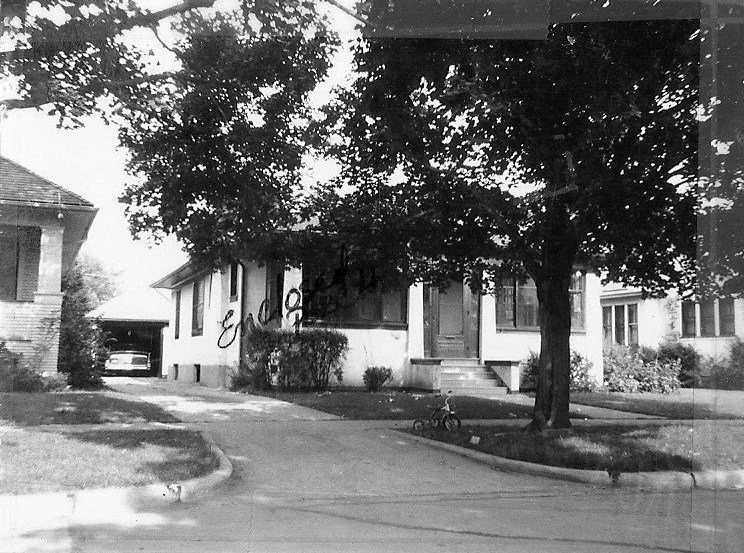 Black-and-white photo of a single-story bungalow with a front porch, large shade tree, driveway to a garage, and a child's tricycle on the lawn.