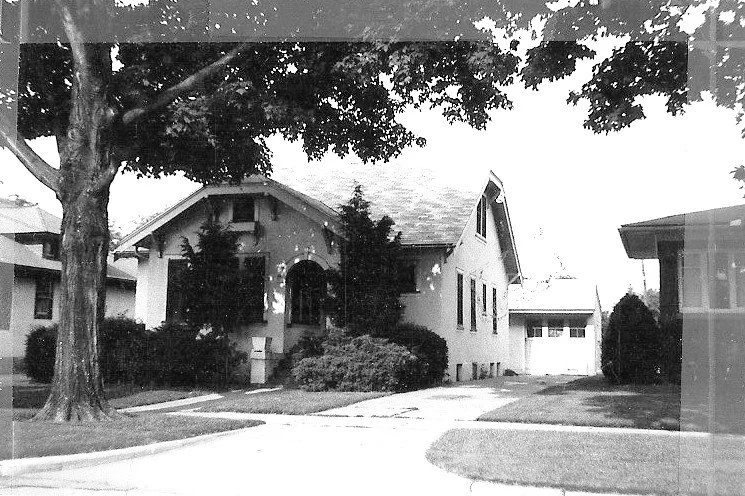 Black-and-white photo of a bungalow-style house with a front porch, large tree, driveway, and detached garage.