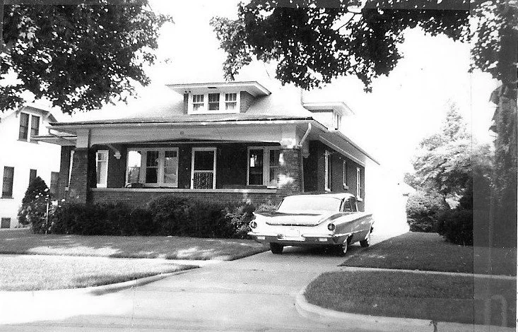 Black-and-white photo of a single-story brick bungalow with a covered front porch and a vintage car parked in the driveway, framed by trees on a residential street.