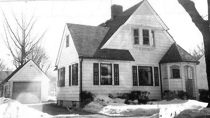 Black-and-white photo of a two-story gabled house with a detached garage and a snow-covered front yard.