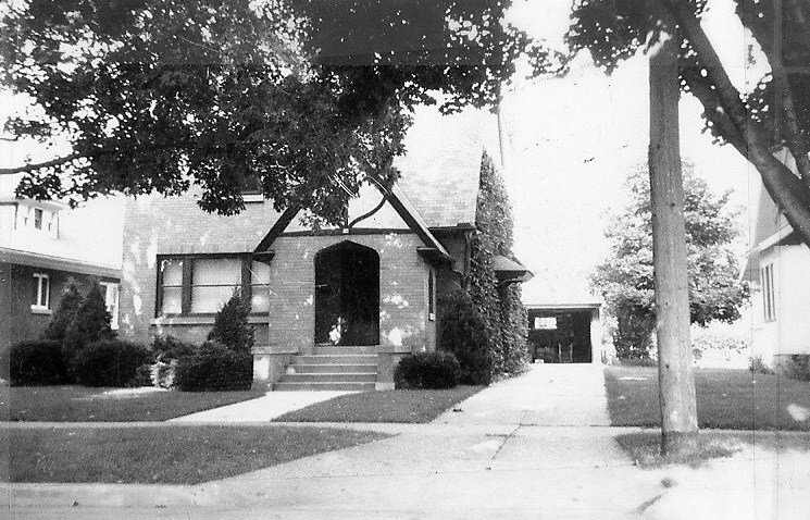 Black-and-white photo of a brick bungalow with an arched front porch, front steps, driveway leading to a detached garage, and large trees.